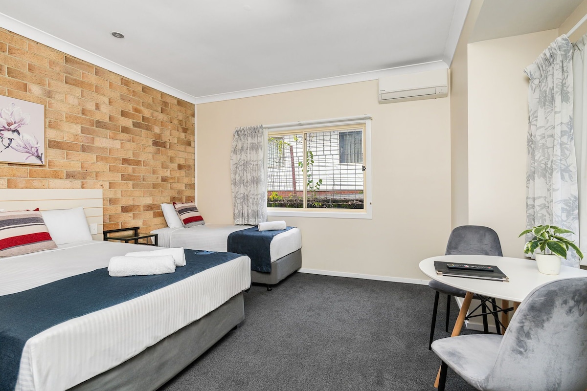 A hotel room with two beds, a small table with a chair, a potted plant, and a window with curtains.