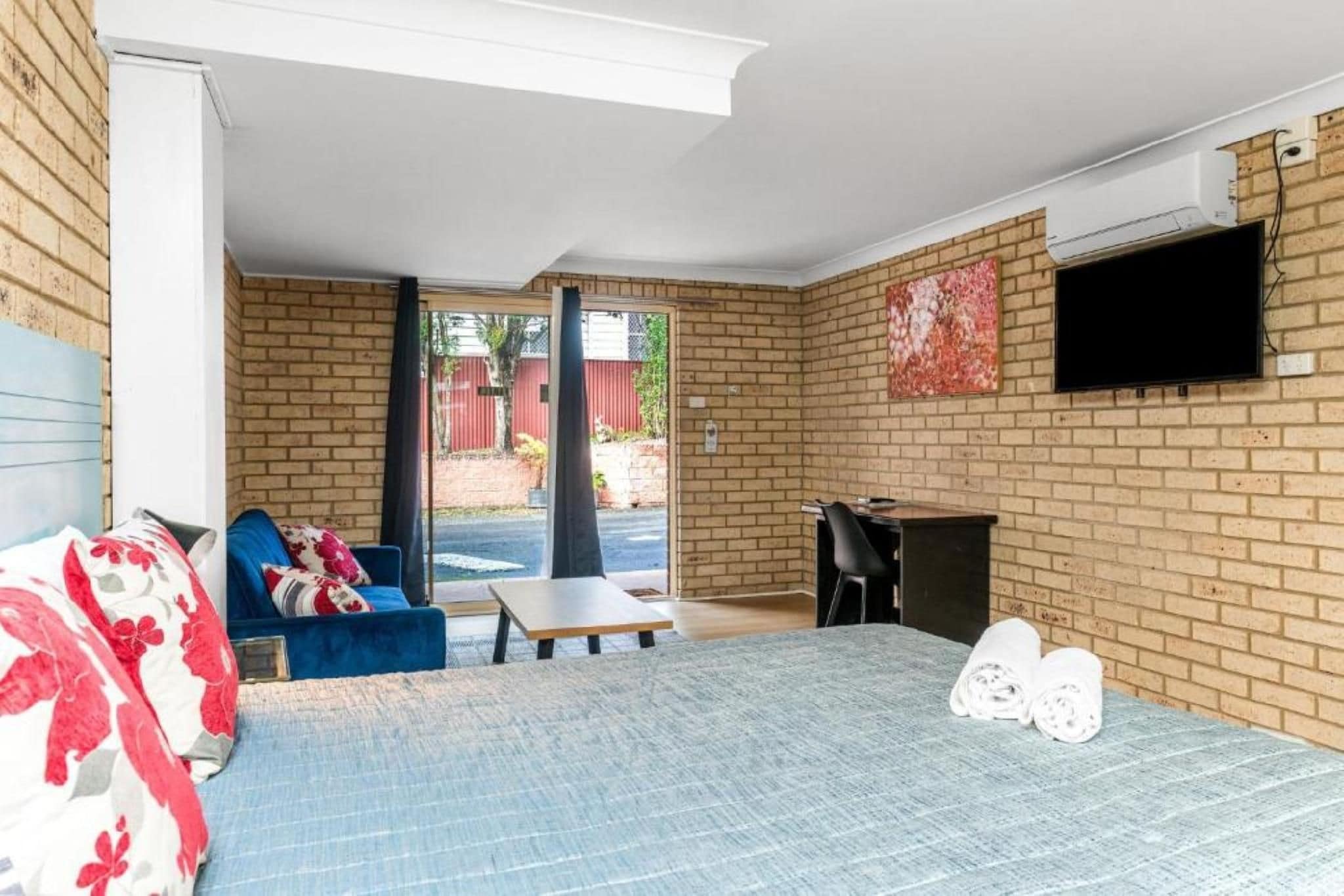 A room with a bed, a blue sofa, a small table, a TV mounted on the wall, and a brick wall.
