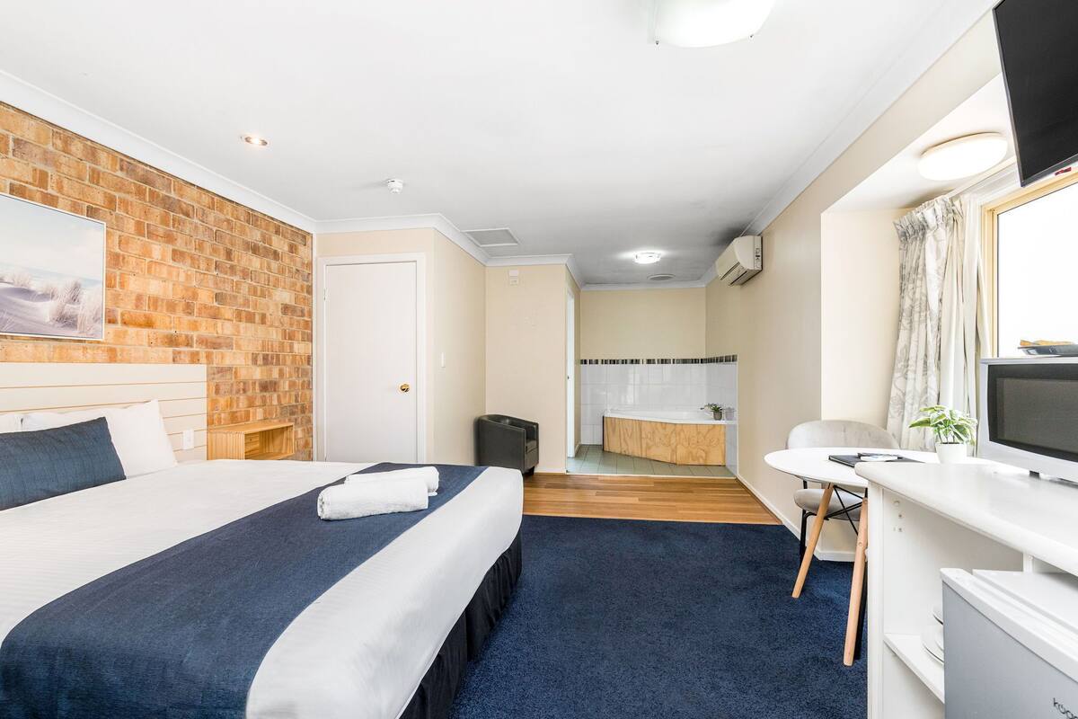Habitación de hotel con una cama grande, un escritorio, una silla, un televisor y un baño con bañera, además de una pared de ladrillo.