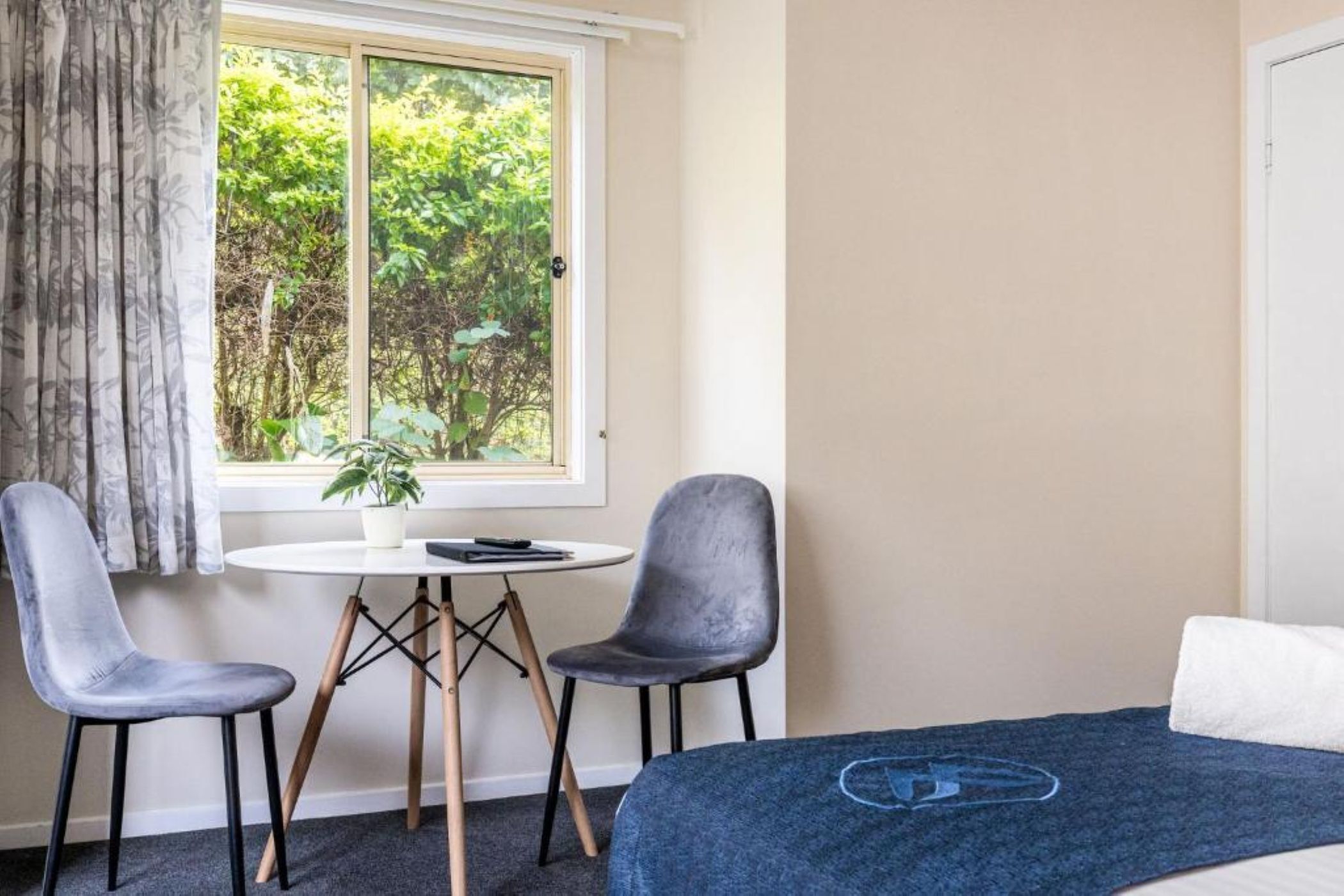 Een kamer met een bed, twee stoelen, een tafeltje met een plant en een raam met uitzicht op groen.