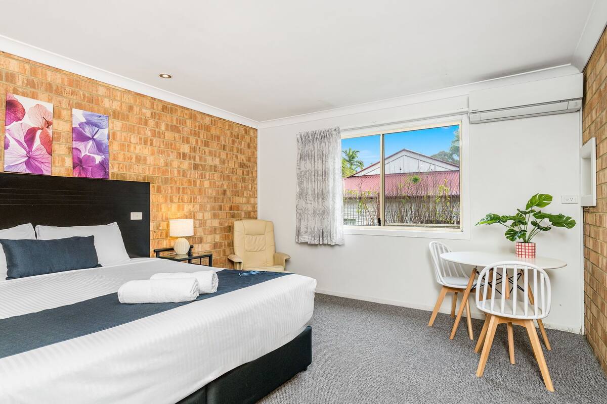 Una habitación de hotel con cama, una mesita con sillas, una ventana con cortinas y una pared de ladrillo.