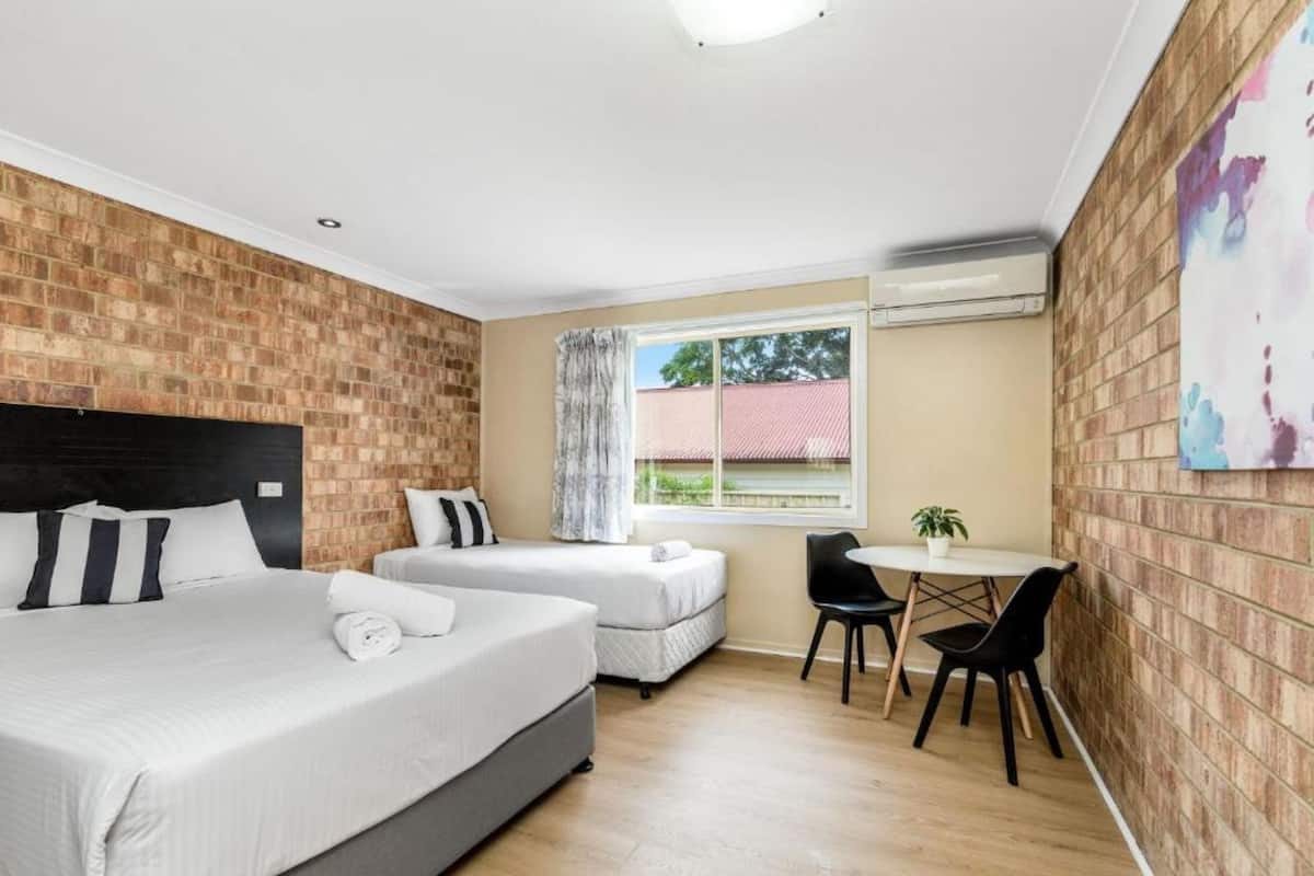 Habitación de hotel con pared de ladrillo, una cama grande, una cama más pequeña, una ventana con vistas, una mesa con sillas y un equipo de aire acondicionado.