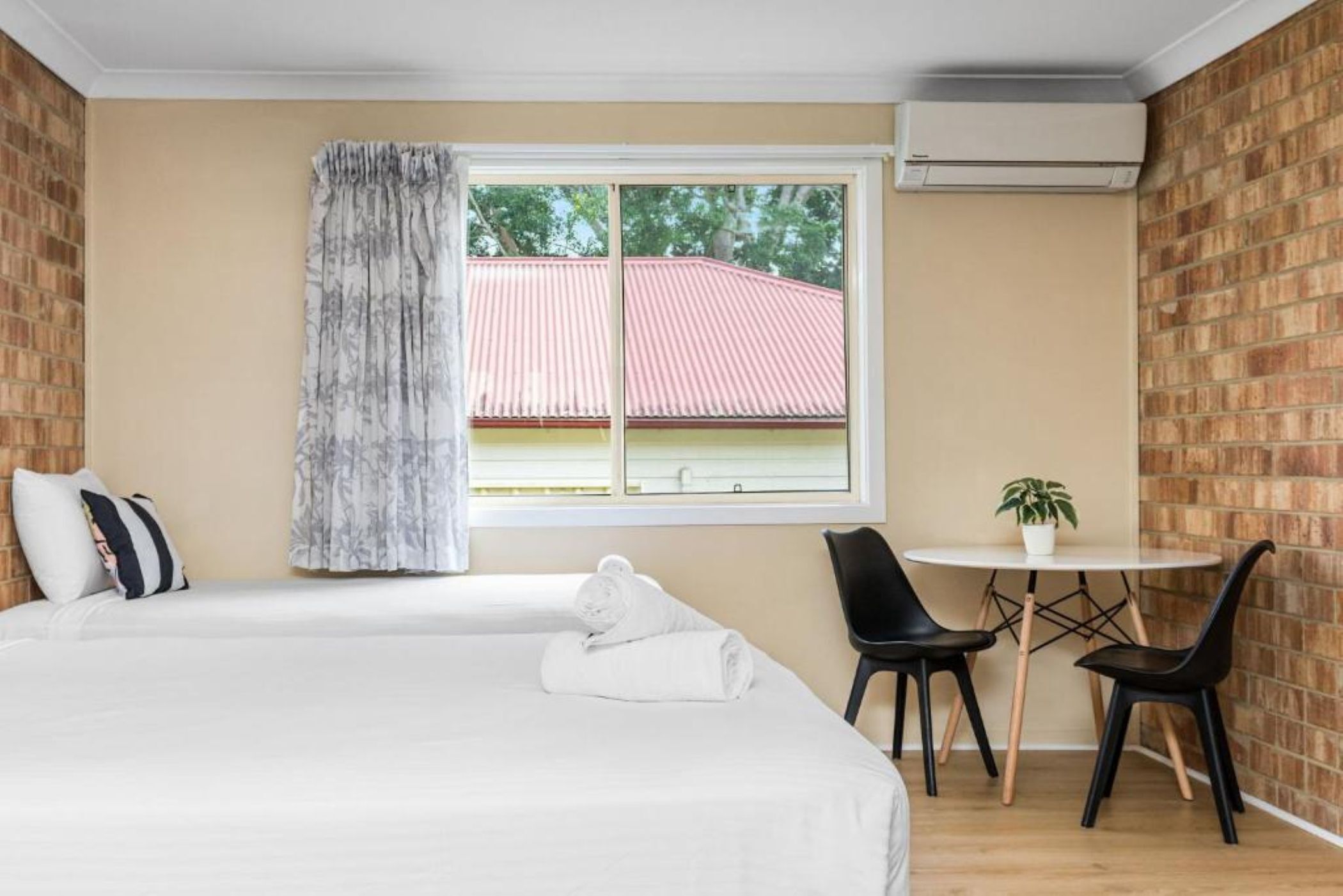 Ein Hotelzimmer mit Bett, Fenster mit Blick auf ein Gebäude mit rotem Dach, einem kleinen Tisch und zwei Stühlen sowie einer Klimaanlage.