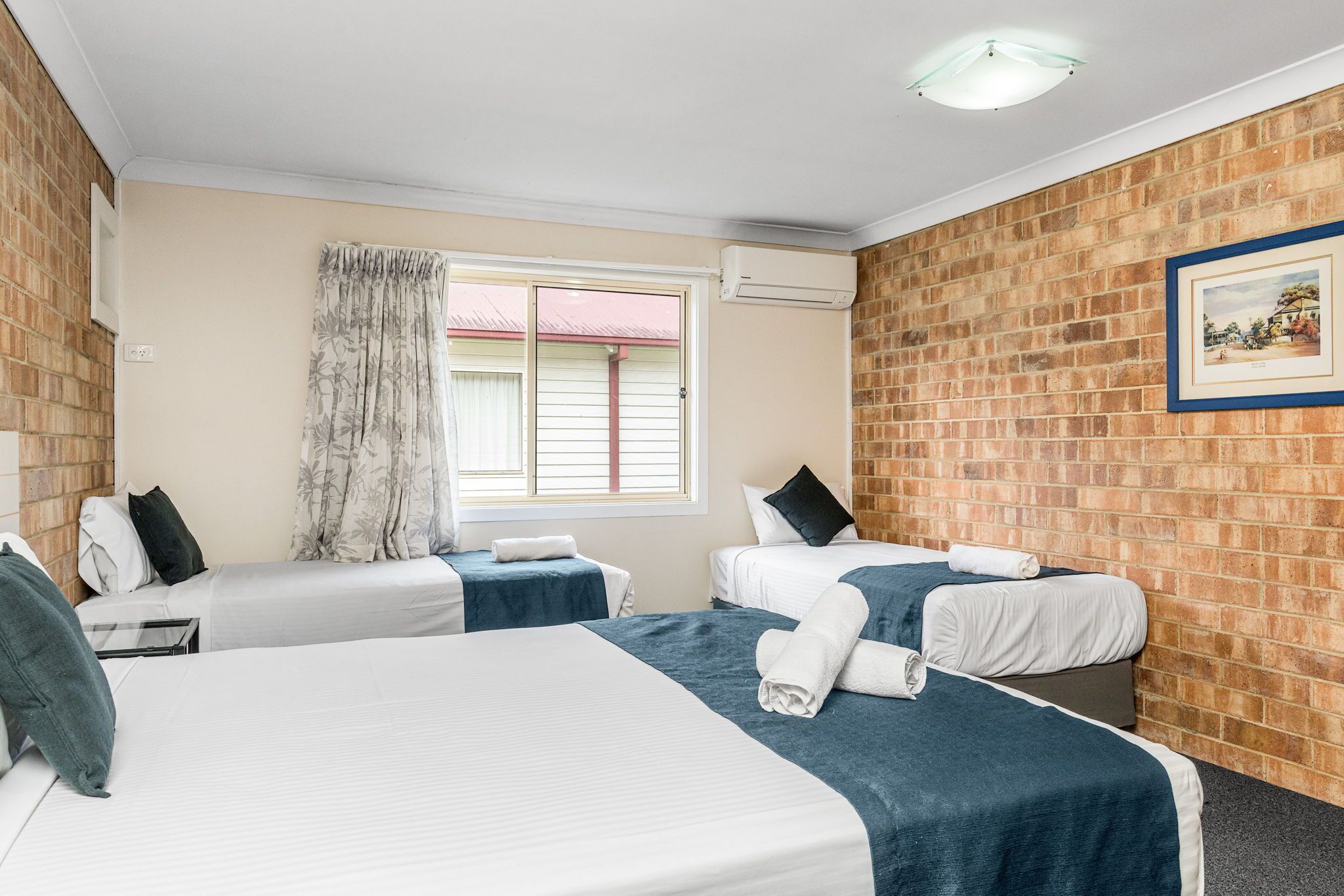 Una habitación de hotel con dos camas individuales, un ventanal con cortinas, una pared de ladrillo y un cuadro enmarcado.