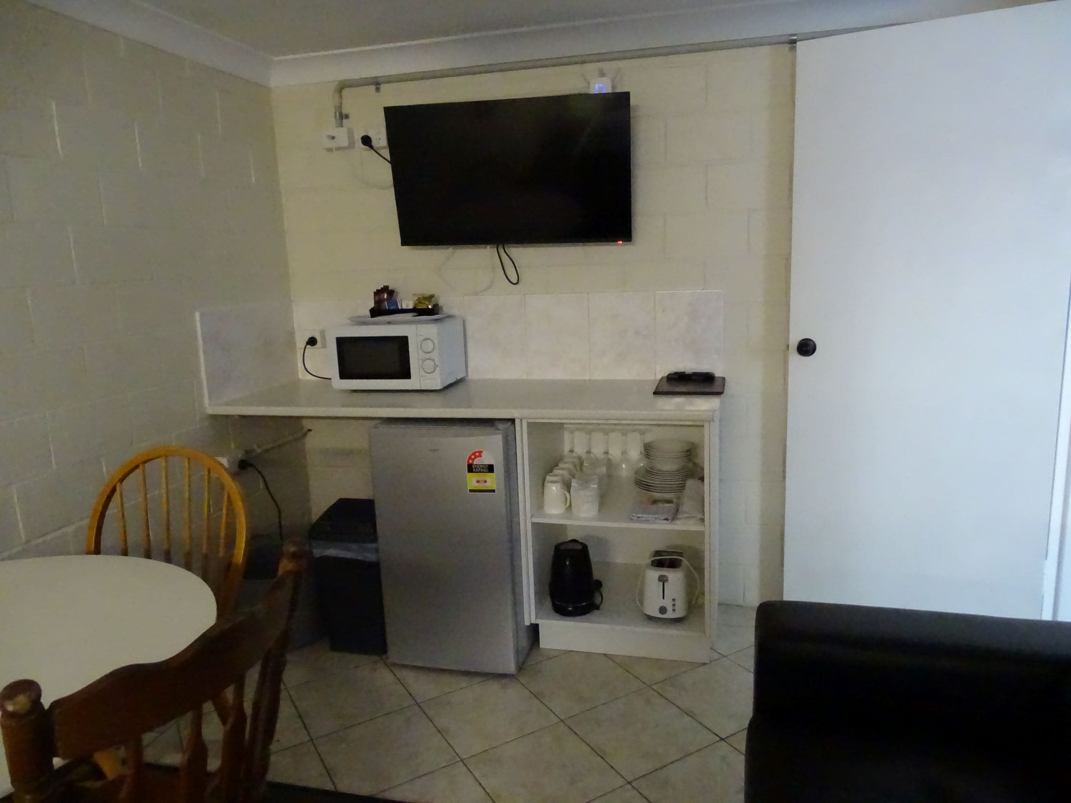 A compact kitchen with a microwave, refrigerator, and a shelf with kitchenware.