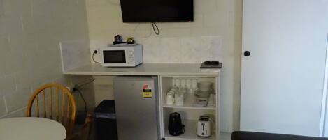 A compact kitchen with a microwave, refrigerator, and a shelf with kitchenware.