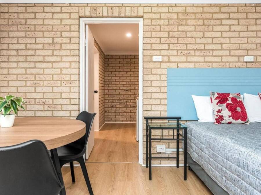 A room with a brick wall, a bed with floral pillows, a table with chairs, and a potted plant.