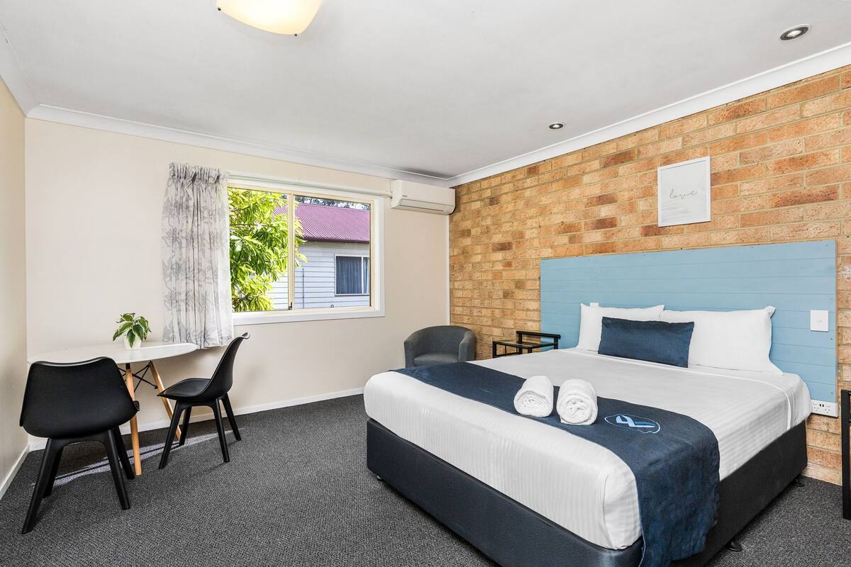 Habitación de hotel con cama, dos sillas, una mesita, ventana con cortinas y pared de ladrillo.
