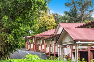 Filas de habitaciones de motel con techo rojo y construcción de ladrillo, con un paseo cubierto y abundante vegetación al fondo.