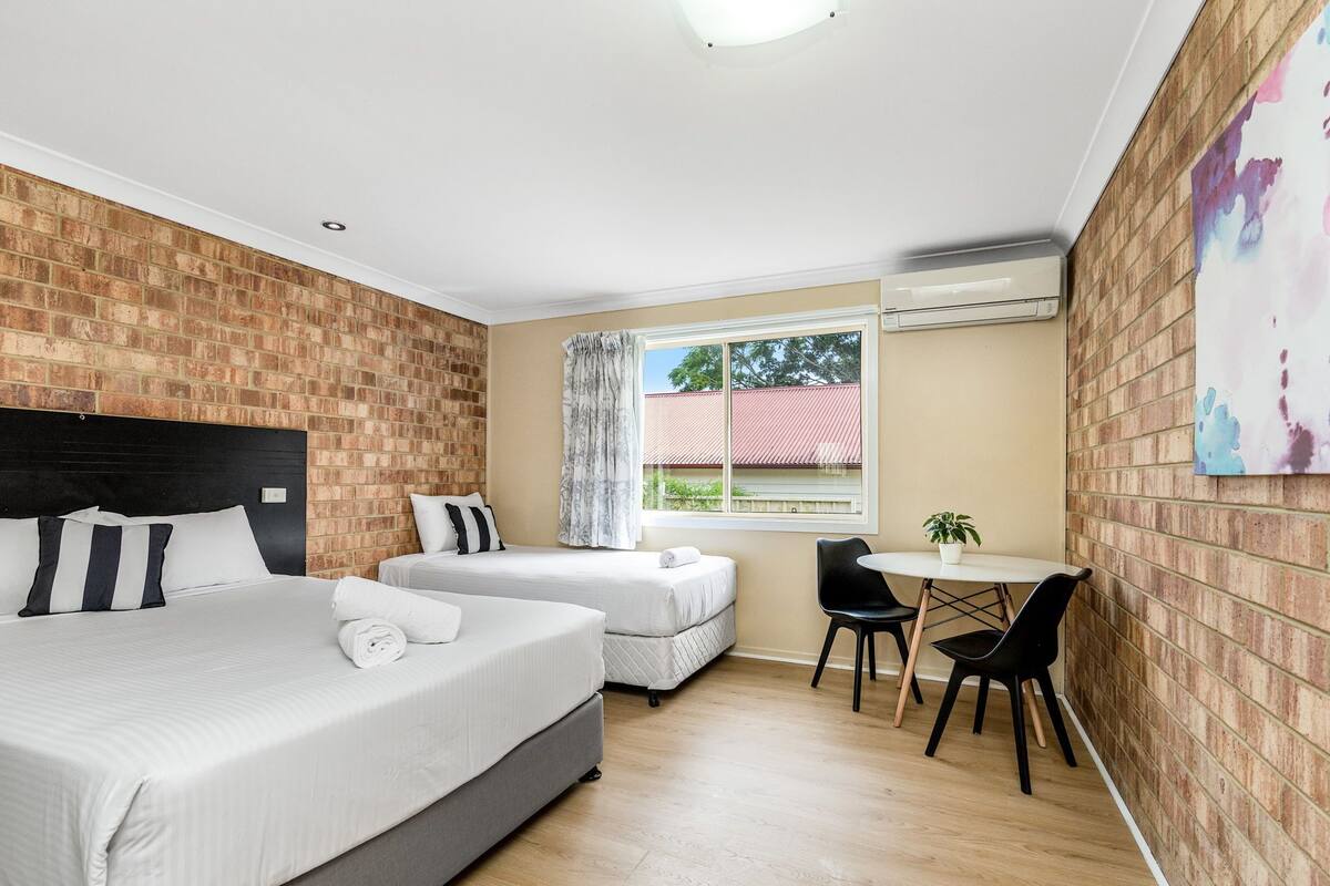 Habitación de hotel con dos camas, una mesa de comedor con sillas, una ventana con cortinas y una pared de ladrillo.