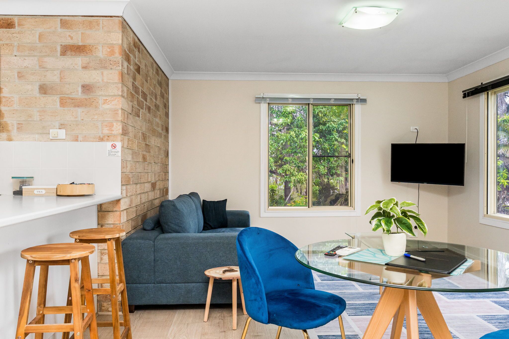 A modern living room with a sofa, a blue chair, a glass table, wooden stools, a potted plant, and a TV mounted on the wall.