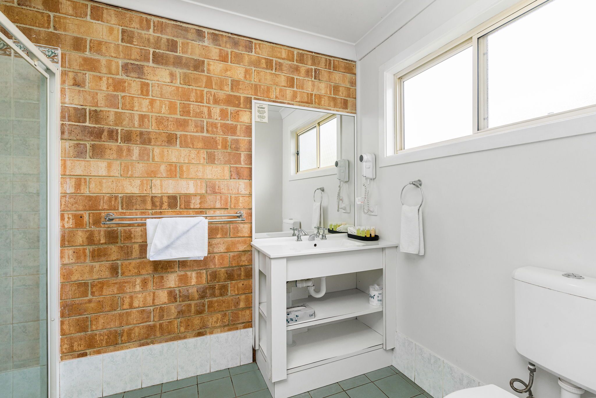Un baño con pared de ladrillo, un lavamanos blanco, un espejo y un inodoro.