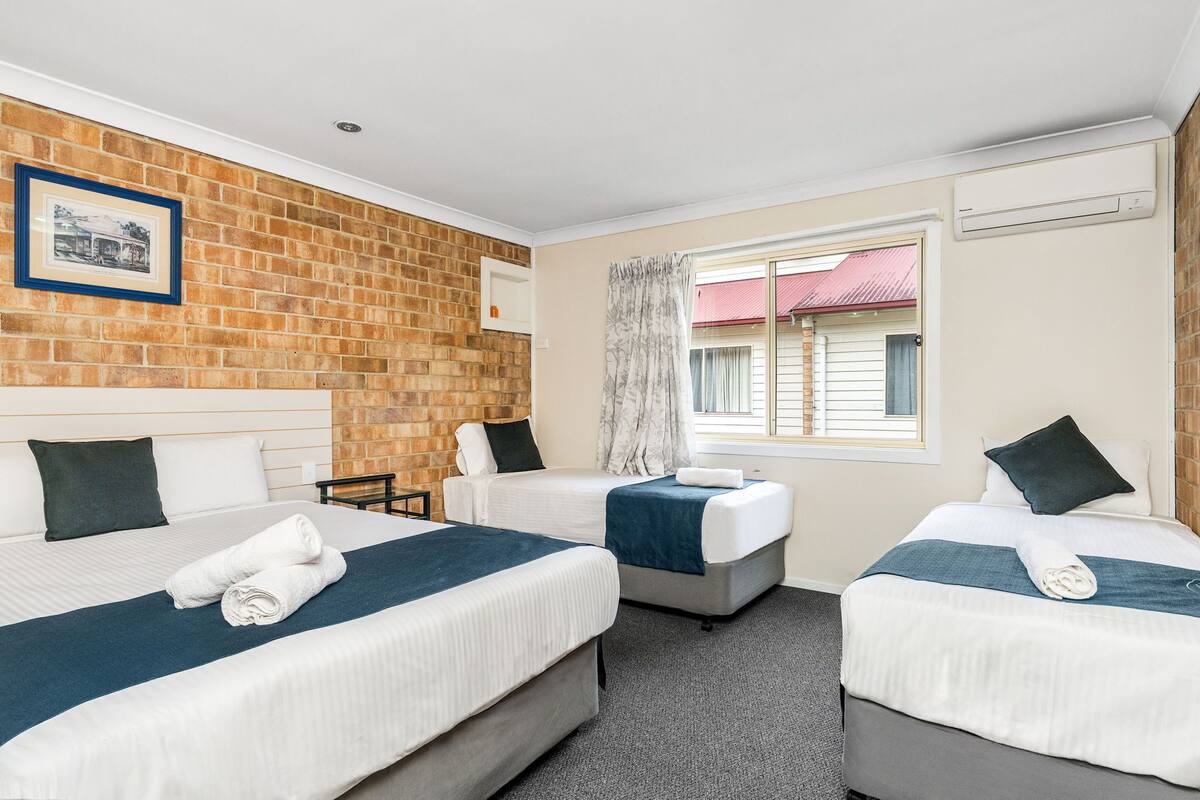 Habitación de hotel con tres camas individuales, una pared de acento de ladrillo, un cuadro enmarcado y una ventana con cortinas.