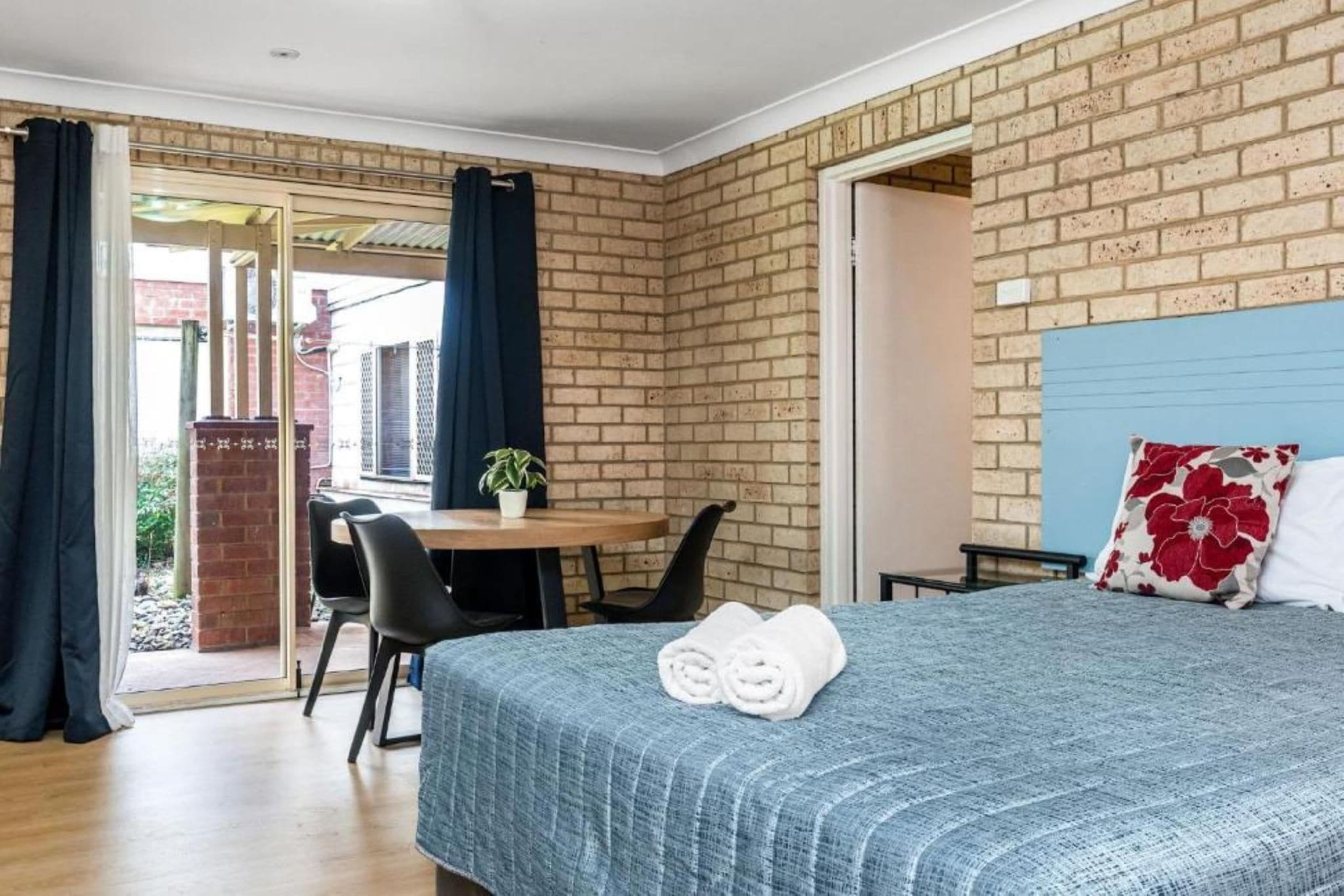 Habitación de hotel con una cama, un escritorio y una silla. Se ve una ventana con cortinas, una pared de ladrillo y un cabecero azul.