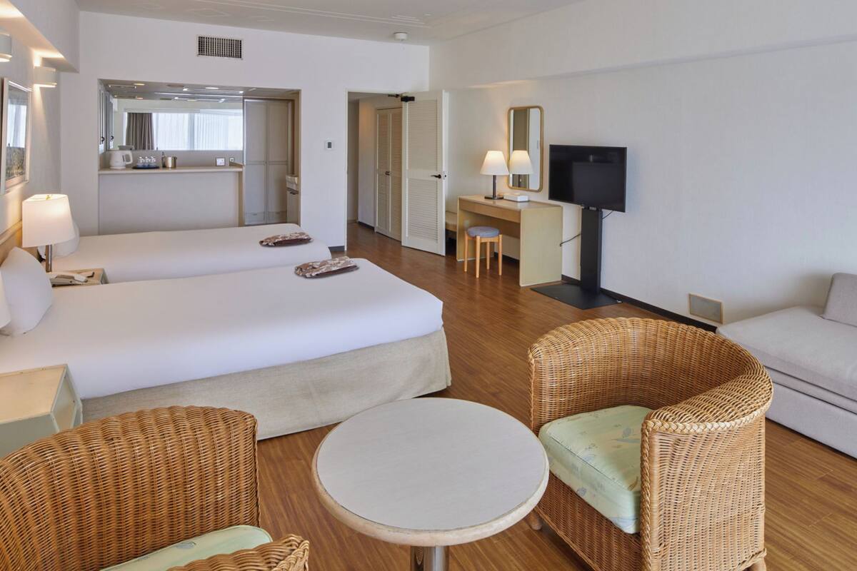 A hotel room with a bed, two wicker chairs, a sofa, a small table, a desk with a TV, and a mirror.