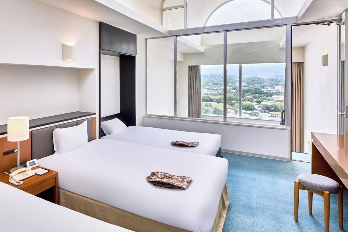 A hotel room with two beds, a desk, a chair, a phone, and a large window with a city view.