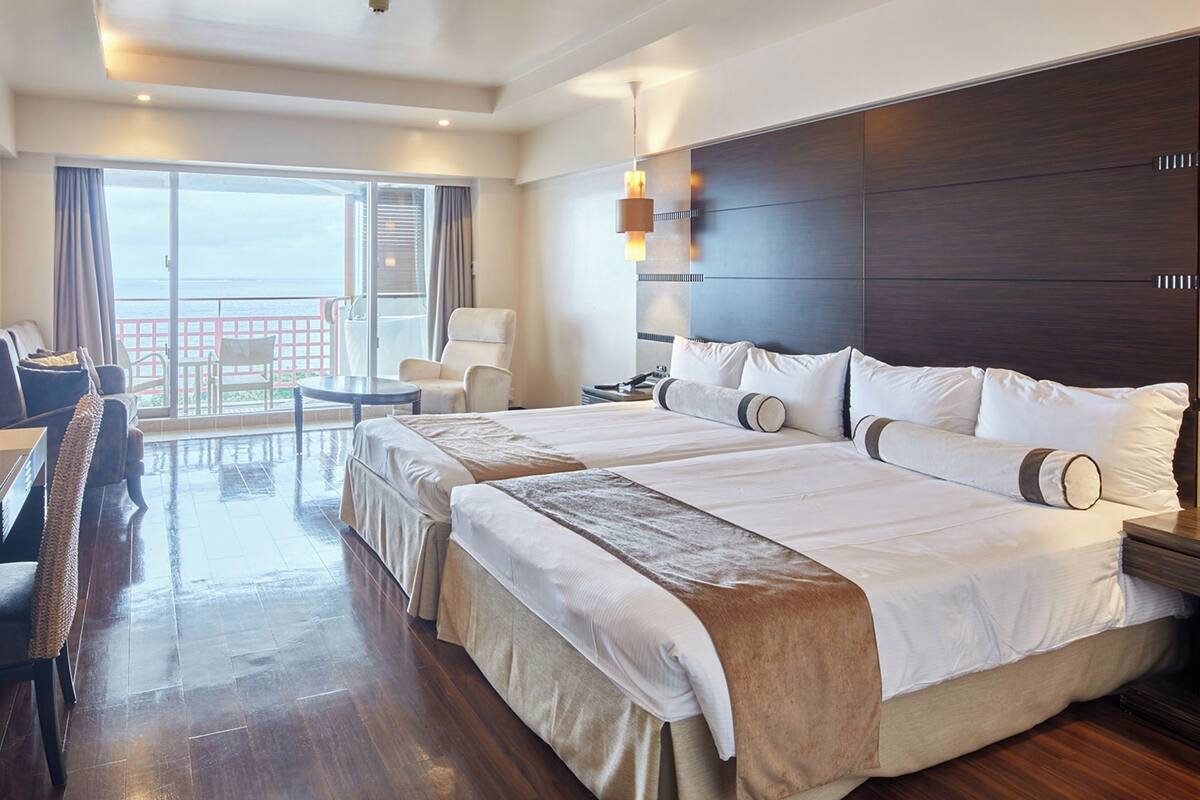 A hotel room with a large bed, a desk, a chair, a small table, and a balcony with a view.