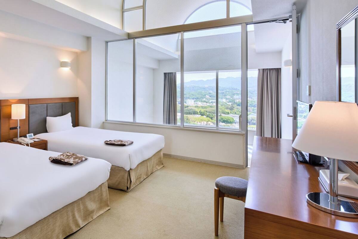 A hotel room with two beds, a desk, a lamp, and a large window offering a city view.