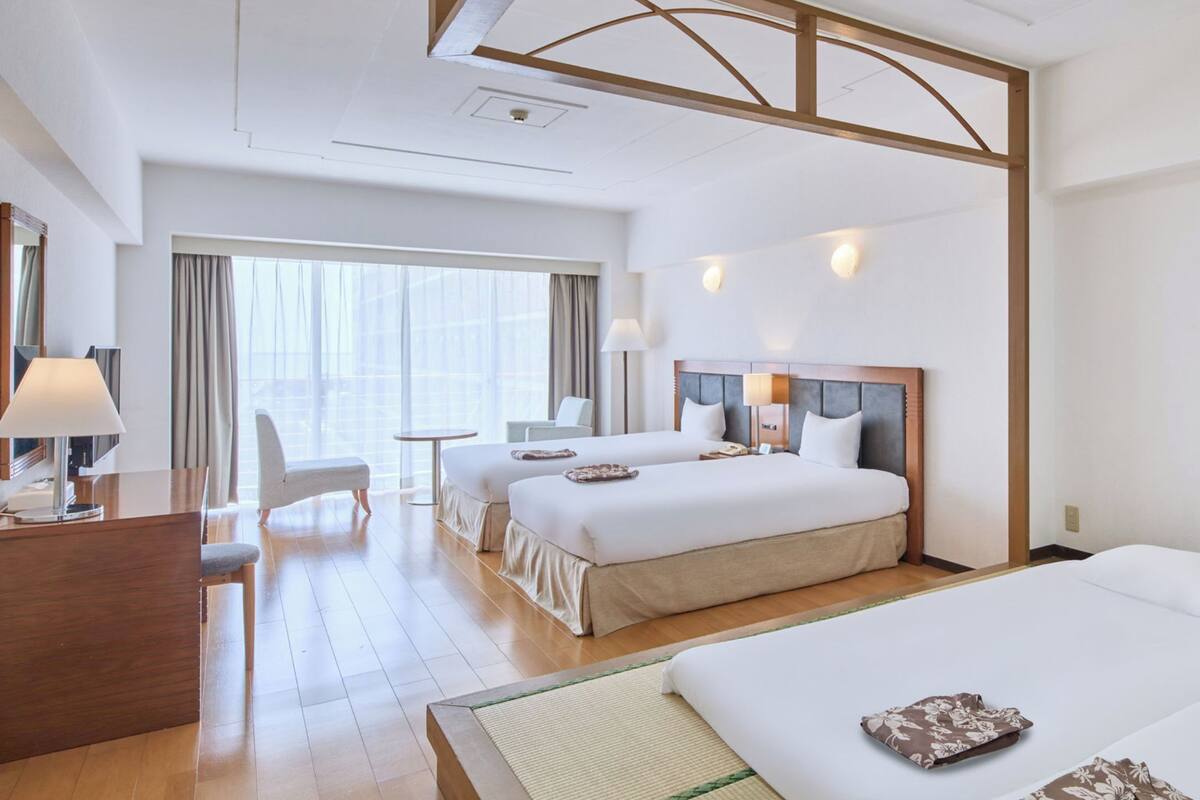 A hotel room with two beds, a desk, a chair, a lamp, and a view of the outdoors.