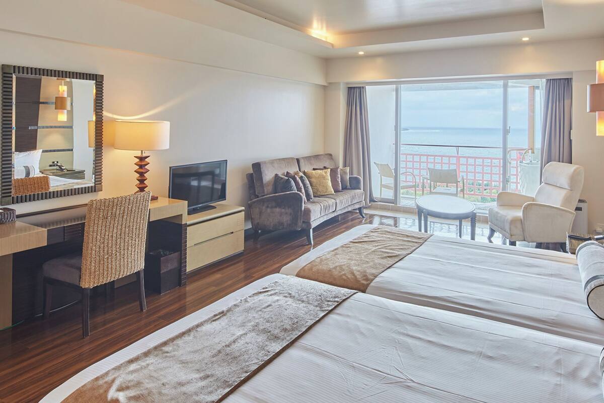 A hotel room with a large bed, a desk with a TV, a chair, a sofa, and a balcony with a view.