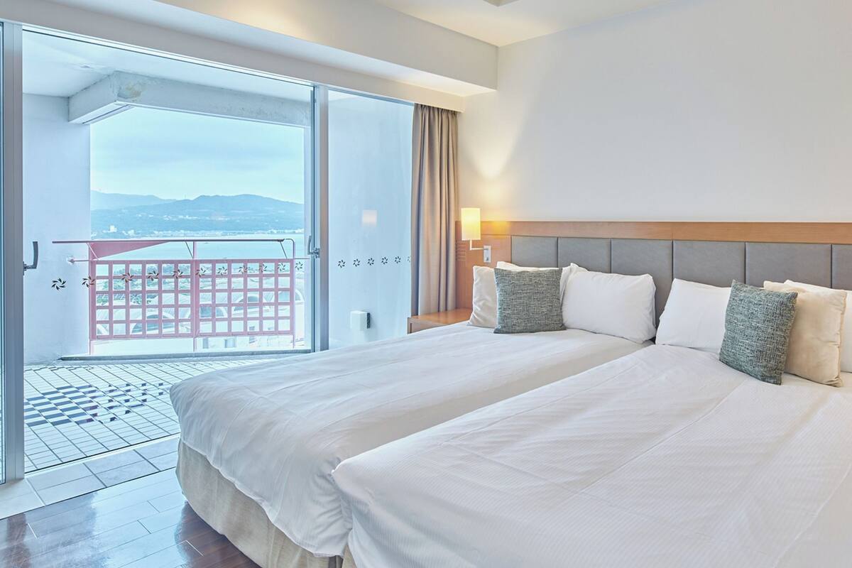 A hotel room with a large bed, two bedside tables, a sliding glass door to a balcony, and a view of the mountains.