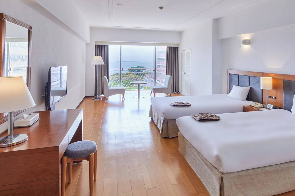 A hotel room with two beds, a desk, a chair, a TV, and a balcony with a view.