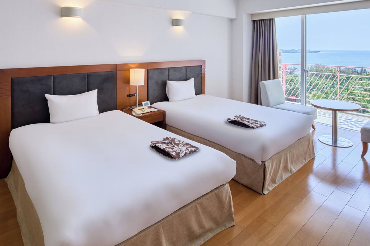 A hotel room with two beds, a small table, a chair, and a balcony with a view of the sea.