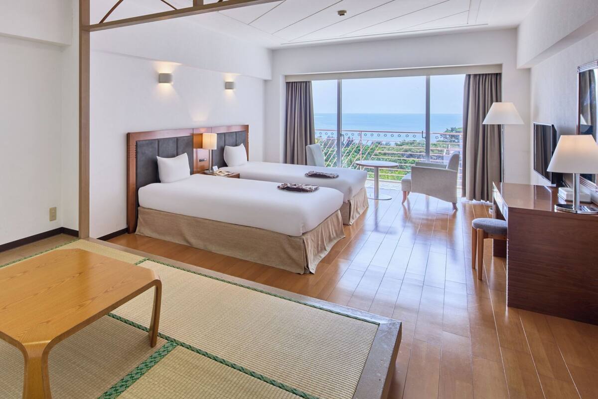 A hotel room with two beds, a desk, a chair, a table, and a view of the sea.