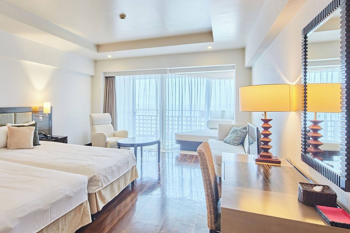 A hotel room with two beds, a sitting area with a chair and table, a large mirror, and a view through the window.