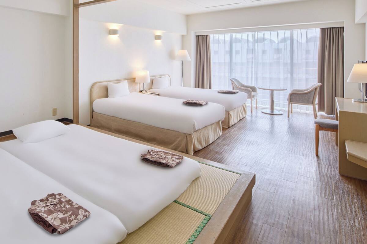 A hotel room with two beds, a desk, a chair, and a view of the city.