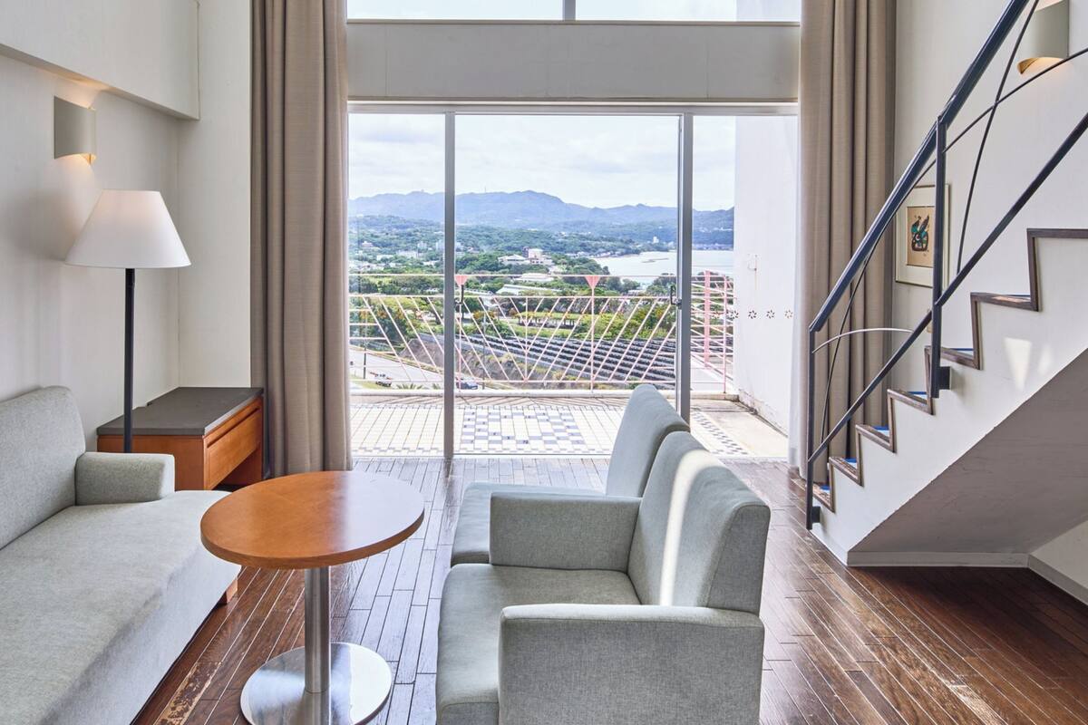 A modern hotel room with a sofa, a small round table, a lamp, and a staircase leading to a balcony with a city view.
