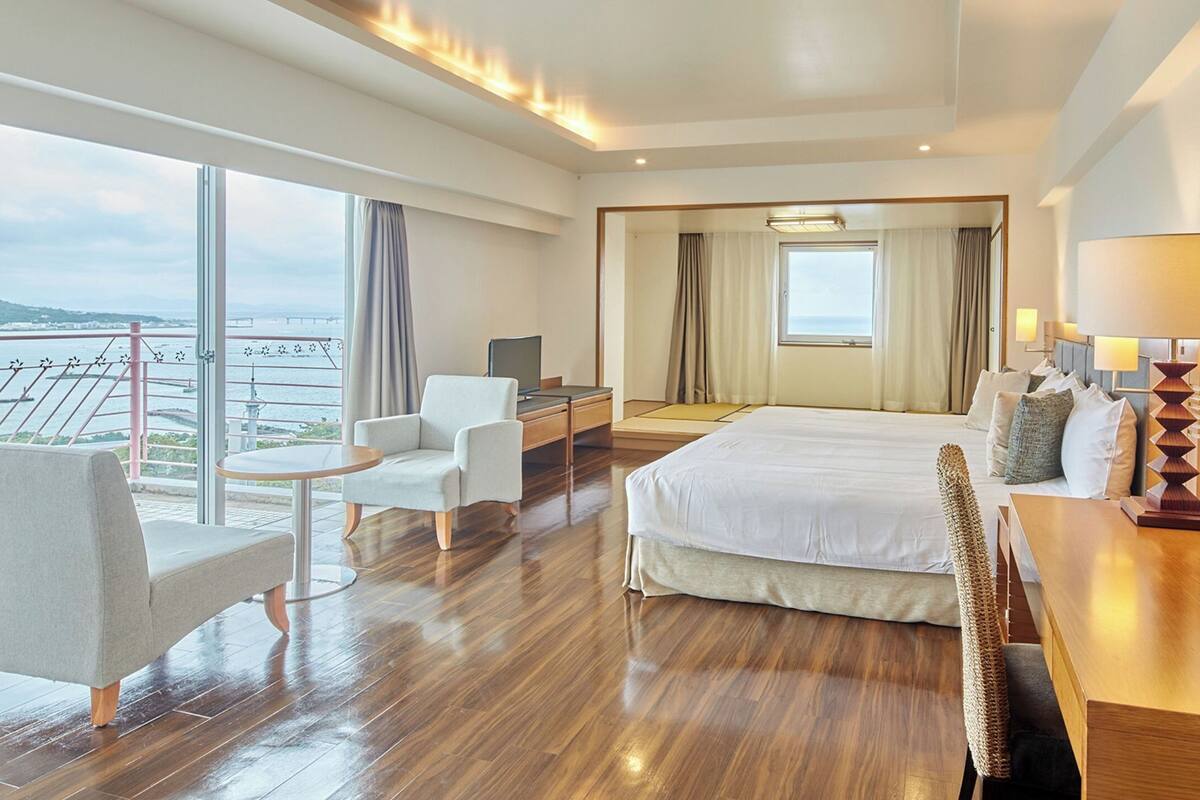 A hotel room with a bed, a desk, a chair, a small table, a mirror, and a balcony with a view of the sea.