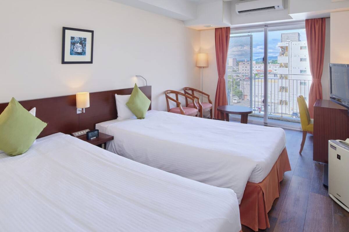Une chambre d’hôtel avec deux lits, un bureau, une chaise, une télévision et un balcon donnant sur la ville.