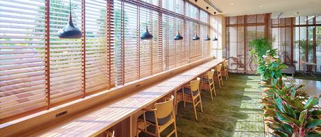 A modern interior with a long wooden counter, multiple chairs, and large windows with blinds.