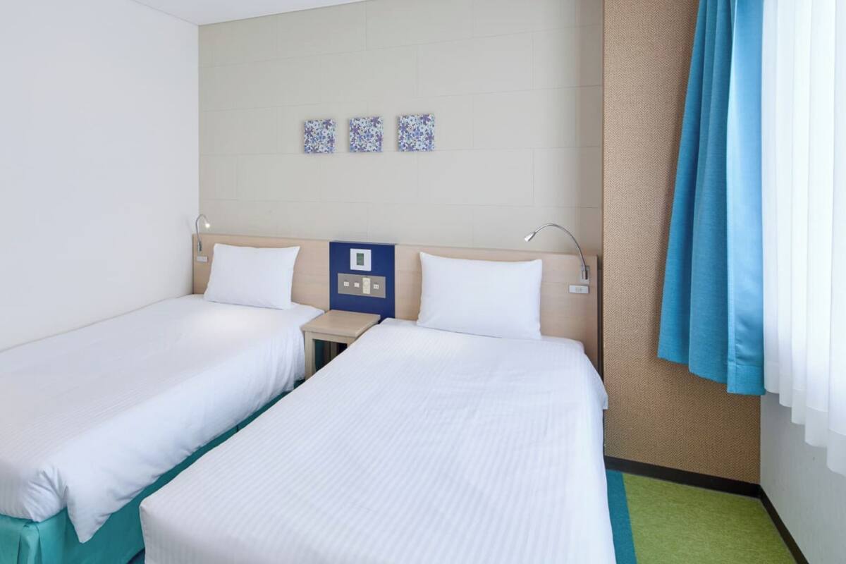 Une chambre d’hôtel avec deux lits simples, une petite table, un panneau de commande fixé au mur et une fenêtre avec des rideaux.