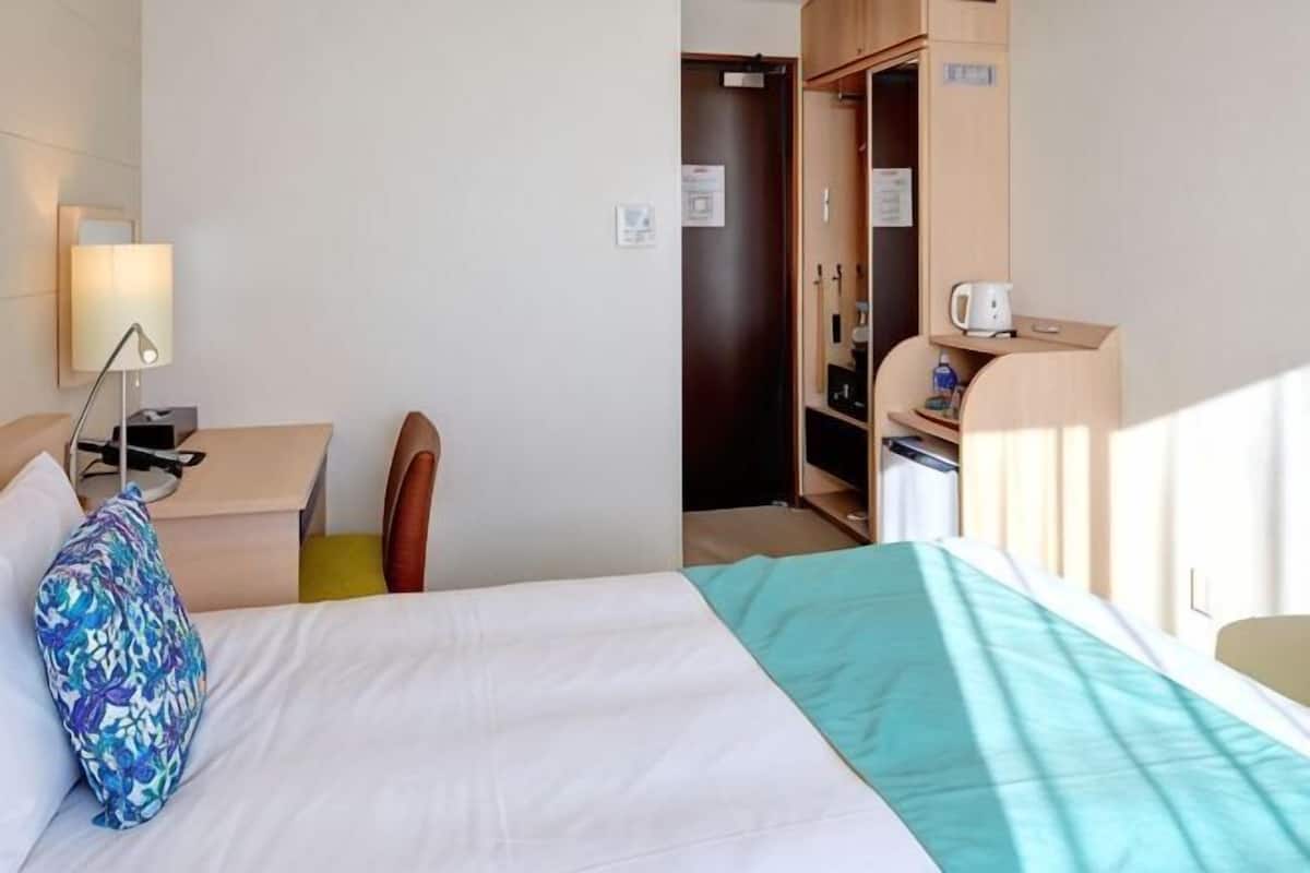 Una habitación de hotel con cama, escritorio, silla, armario y una pequeña kitchenette.