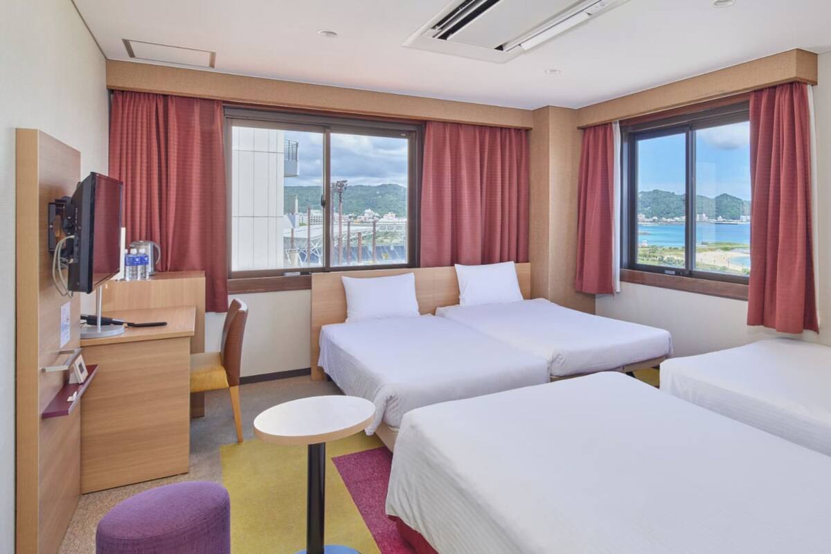 Une chambre d’hôtel avec deux lits, un bureau avec un ordinateur, une télévision et de grandes fenêtres avec des rideaux.