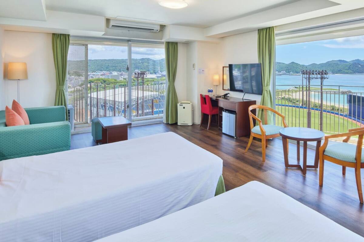 Une chambre d’hôtel avec deux lits, un bureau, une chaise et une télévision. La chambre dispose de grandes fenêtres donnant sur un plan d’eau et des collines.