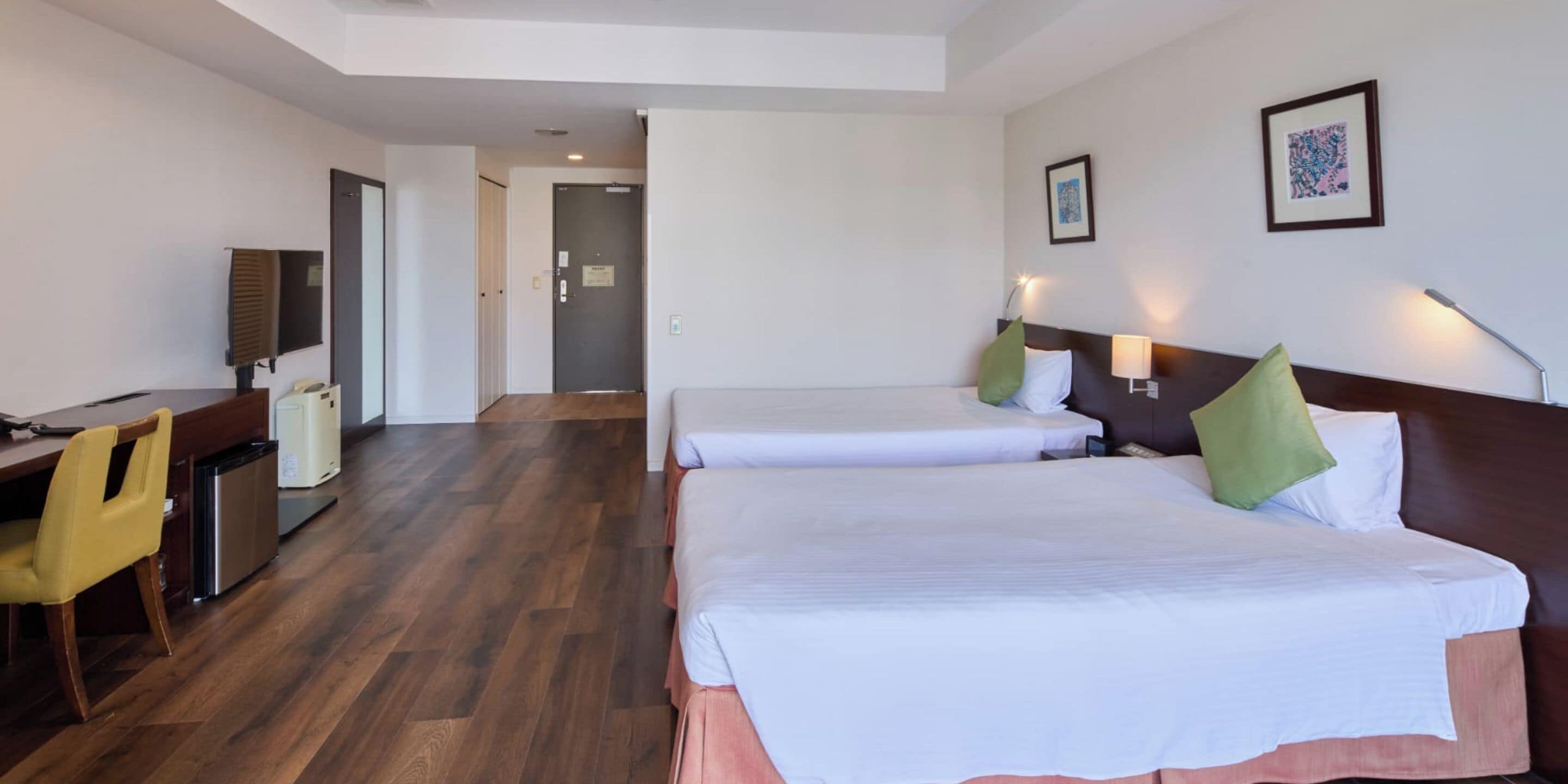Habitación de hotel con dos camas, un escritorio, una silla, un refrigerador pequeño y una puerta que conduce a otra habitación.