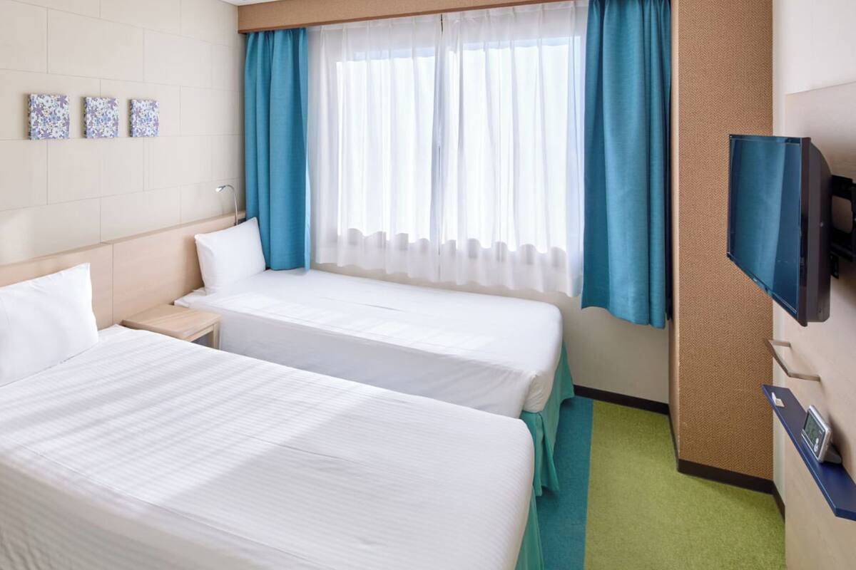 Une chambre d’hôtel avec deux lits, une télévision, un bureau avec une lampe, un miroir et une fenêtre avec des rideaux.