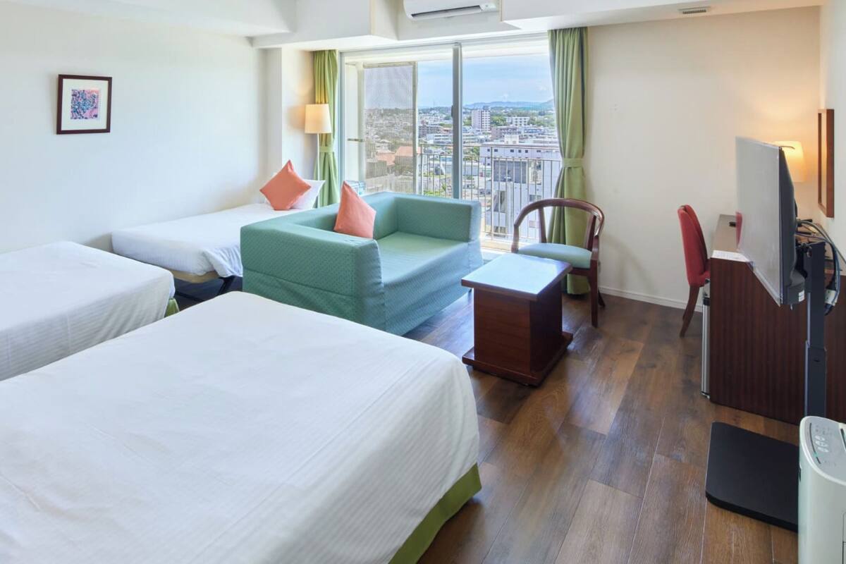 Une chambre d’hôtel avec deux lits simples, un canapé, un bureau, une chaise, une télévision et une fenêtre donnant sur la ville.