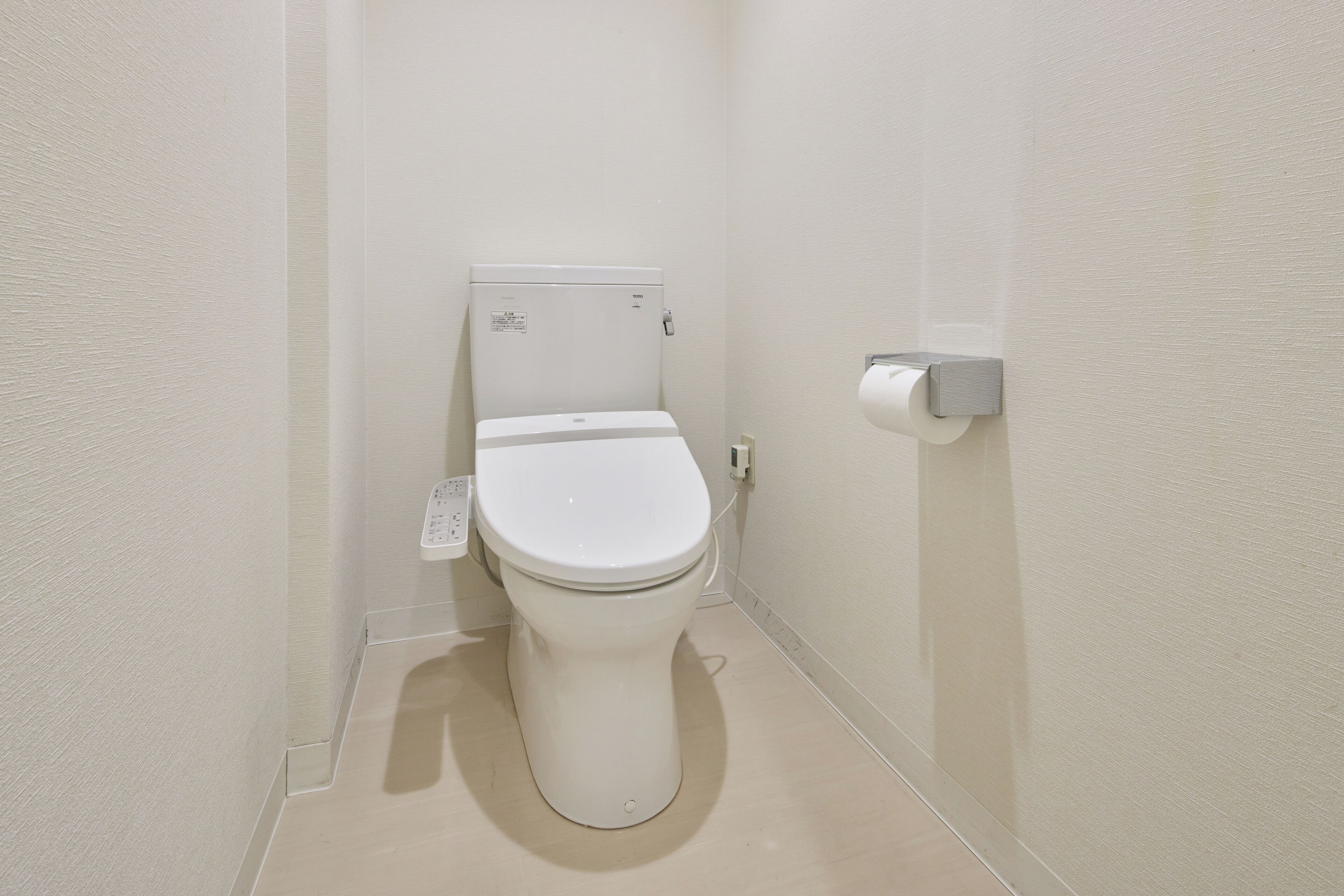 A small, white bathroom with a toilet, a paper towel dispenser, and a wall-mounted electrical outlet.