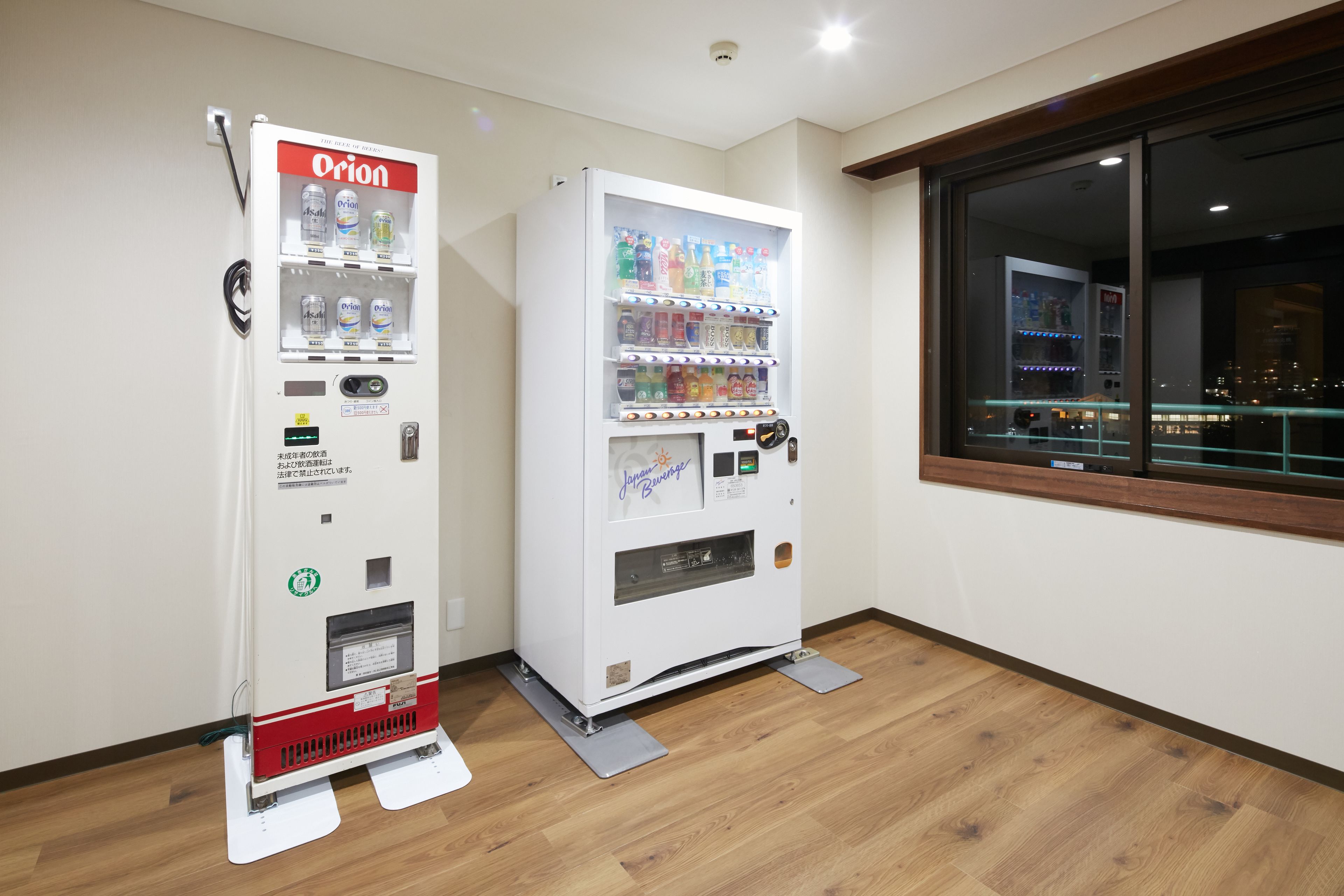 Two vending machines in a room with wooden flooring and a window with a view of the city at night.