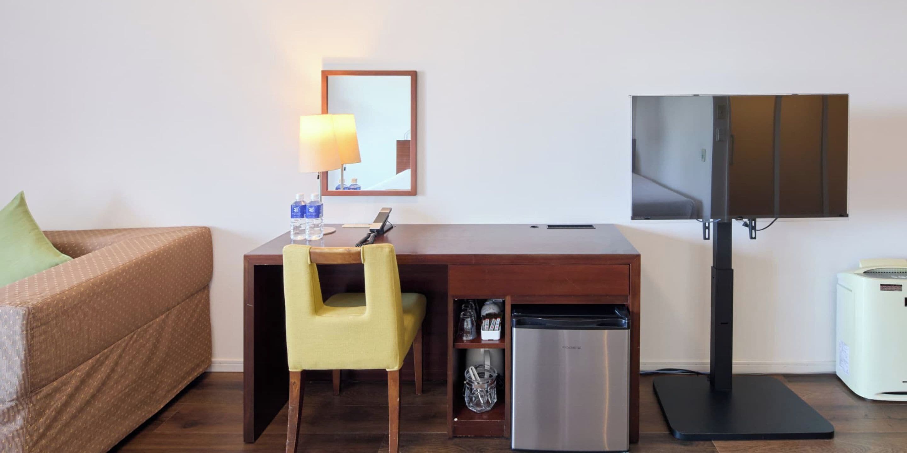 A compact hotel room with a wooden desk, a yellow chair, a small refrigerator, a television, and a sofa.