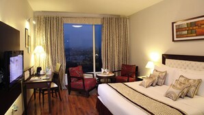 A hotel room with a large bed, two armchairs, a small table, a TV, and a window with curtains. - TreeHouse Queens Pearl (Gurugram)