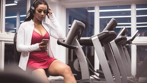 Een persoon zit op een fitnessapparaat, draagt een witte cardigan en een rode jurk, met een telefoon in de hand.