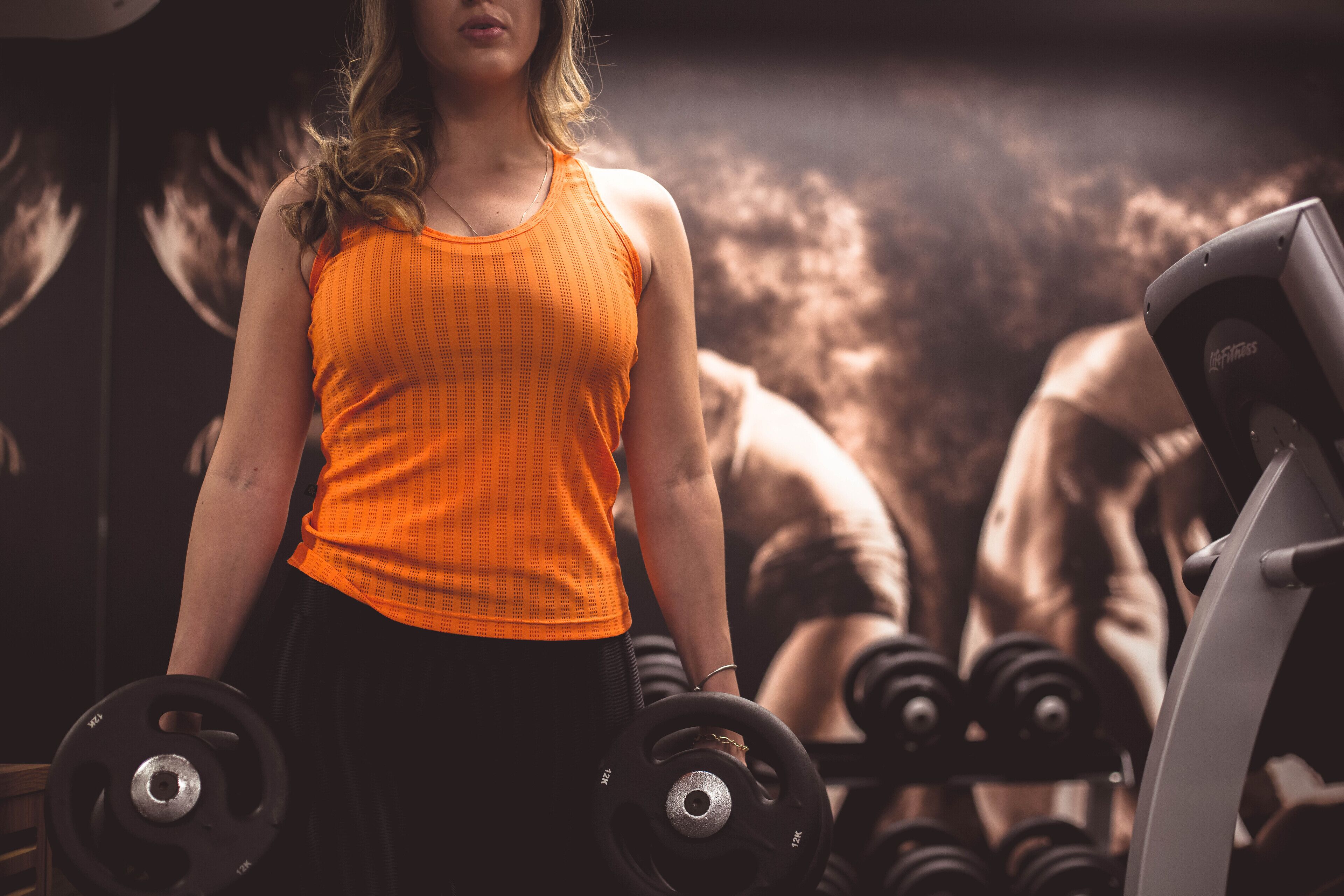 Uma pessoa segurando halteres numa academia, com equipamento de exercícios ao fundo.