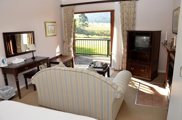 Una habitación de hotel con una cama, un sofá a rayas, un escritorio con una lámpara, un televisor y un balcón con vistas al campo.