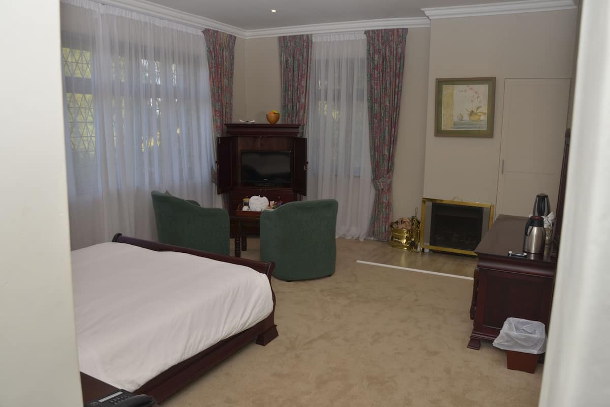 Habitación de hotel con cama, dos sillones verdes, un mueble de televisión de madera, chimenea y una pequeña mesa con un jarrón.