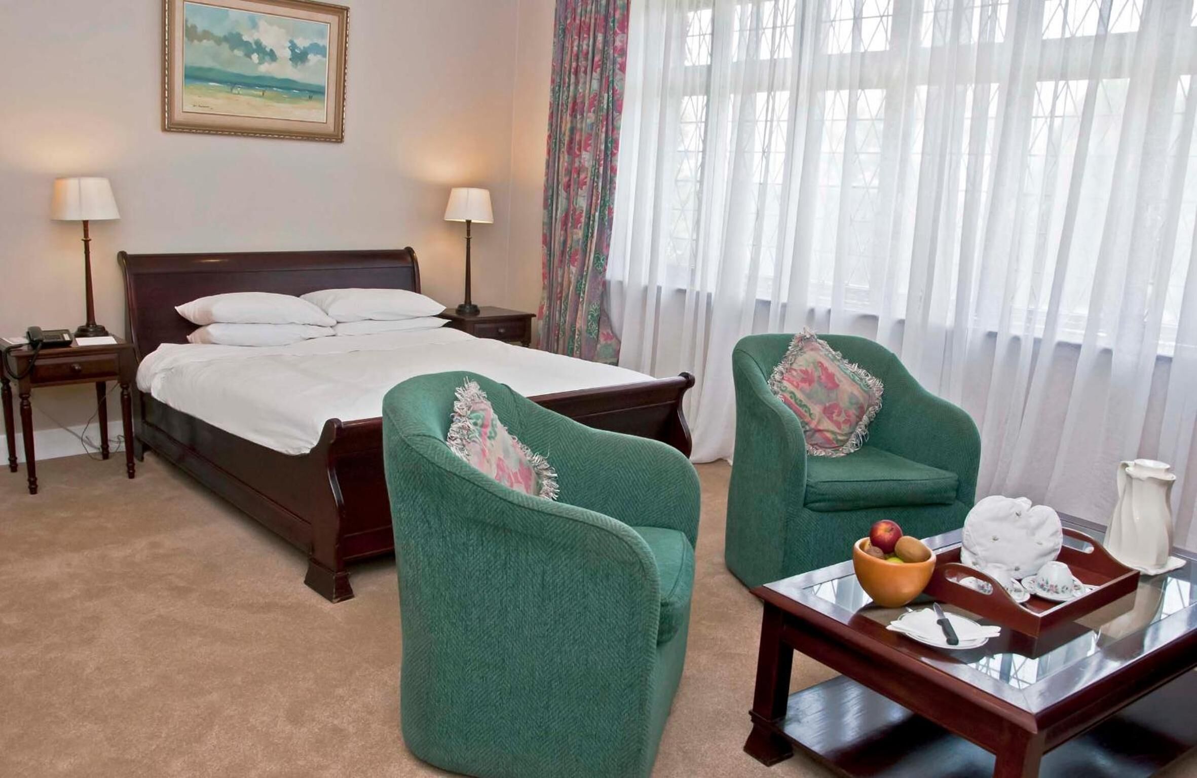 Un dormitorio con una cama grande, dos sillones verdes, una mesita con frutero y tetera, y un cuadro en la pared.